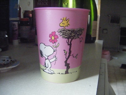 A pink plastic cup with cartoon drawings featuring a dog holding a flower and a bird perched on a nest. The background includes various household items such as bottles and containers.