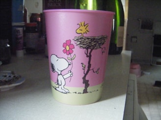A pink plastic cup with cartoon drawings featuring a dog holding a flower and a bird perched on a nest. The background includes various household items such as bottles and containers.