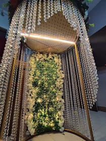 An elaborate decorative piece featuring cascading crystal bead drapes surrounding a central section adorned with lush greenery and white flowers. Warm lights illuminate the arrangement, creating a luxurious and elegant atmosphere. The structure is supported by a metallic frame with a plush, studded ceiling.