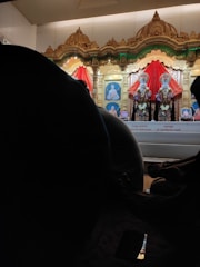 An ornate altar features two richly adorned statues draped in vibrant red fabric, set against a backdrop of intricately carved gold arches. Beneath the statues, photographs are visible, and silhouettes of seated individuals are visible in the foreground. A phone screen reflects some light, adding a touch of modernity to the scene.