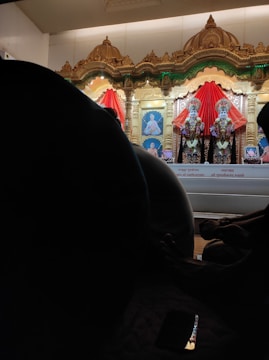 An ornate altar features two richly adorned statues draped in vibrant red fabric, set against a backdrop of intricately carved gold arches. Beneath the statues, photographs are visible, and silhouettes of seated individuals are visible in the foreground. A phone screen reflects some light, adding a touch of modernity to the scene.