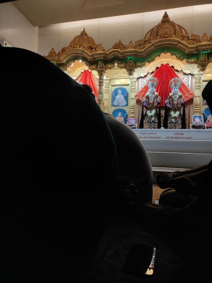 An ornate altar features two richly adorned statues draped in vibrant red fabric, set against a backdrop of intricately carved gold arches. Beneath the statues, photographs are visible, and silhouettes of seated individuals are visible in the foreground. A phone screen reflects some light, adding a touch of modernity to the scene.