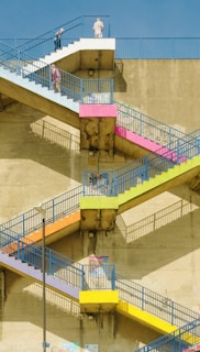 A series of outdoor concrete stairs is painted in bright, vibrant colors including blue, pink, orange, and yellow. The stairs form a zigzag pattern against the wall of a large building. People are walking on the stairs under a clear blue sky. The shadows of the railings create an interesting geometric pattern on the wall.