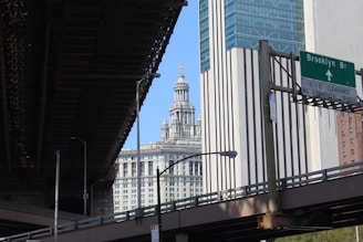 An urban scene showing a highway overpass with a green road sign directing to the Brooklyn Bridge. In the background, an ornate and historical building with intricate architecture is visible against a clear blue sky. Modern skyscrapers with a mix of glass and stone facades surround the area.