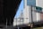 An urban scene showing a highway overpass with a green road sign directing to the Brooklyn Bridge. In the background, an ornate and historical building with intricate architecture is visible against a clear blue sky. Modern skyscrapers with a mix of glass and stone facades surround the area.