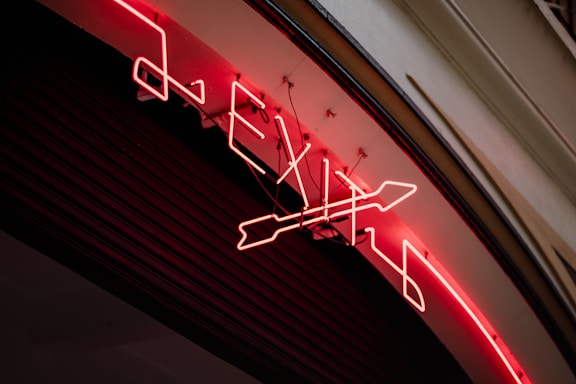 A neon sign displaying the word 'EXIT' along with an arrow symbol is mounted on a curved surface. The neon lights are bright red, illuminating the sign against a dark background. The sign appears to be attached to a building wall, with visible wiring and mounts securing it in place.