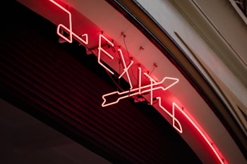 A neon sign displaying the word 'EXIT' along with an arrow symbol is mounted on a curved surface. The neon lights are bright red, illuminating the sign against a dark background. The sign appears to be attached to a building wall, with visible wiring and mounts securing it in place.