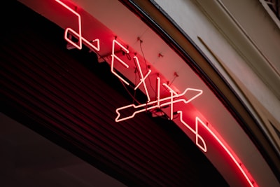 A neon sign displaying the word 'EXIT' along with an arrow symbol is mounted on a curved surface. The neon lights are bright red, illuminating the sign against a dark background. The sign appears to be attached to a building wall, with visible wiring and mounts securing it in place.