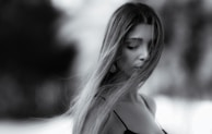 A black and white portrait of a woman with long hair partially covering her face and eyes closed. She appears serene and introspective, wearing a delicate strap on her shoulder. The background is softly blurred, emphasizing her features.