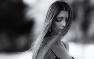 A black and white portrait of a woman with long hair partially covering her face and eyes closed. She appears serene and introspective, wearing a delicate strap on her shoulder. The background is softly blurred, emphasizing her features.