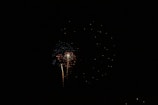Bright fireworks illuminate the dark night sky with bursts of red and white sparks. A trail of light leads upward, suggesting the launch of the firework.