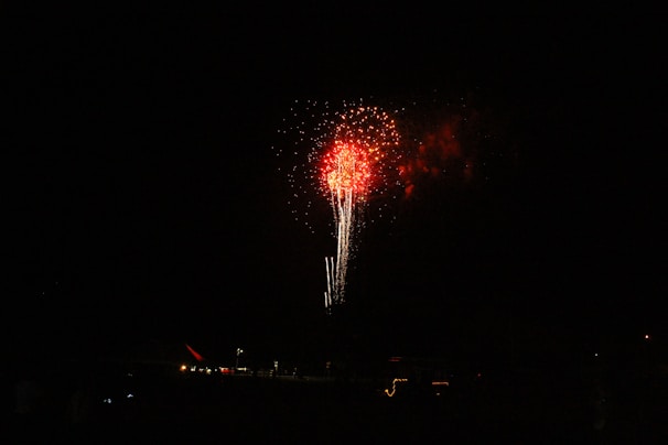 Bright red fireworks explode in the night sky with trails of light below the burst. The surrounding area is dark, with faint outlines of structures and small lights at the bottom, suggesting a distant display.