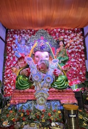 A colorful and ornate display featuring a large statue of the Hindu deity Ganesha, adorned with floral decorations and a garland made of currency notes. The statue is set against a vibrant backdrop of red and white flowers. Flanking Ganesha are detailed images of other deities. The entire setup is richly decorated, giving it a festive and sacred appearance.