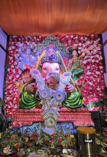 A colorful and ornate display featuring a large statue of the Hindu deity Ganesha, adorned with floral decorations and a garland made of currency notes. The statue is set against a vibrant backdrop of red and white flowers. Flanking Ganesha are detailed images of other deities. The entire setup is richly decorated, giving it a festive and sacred appearance.