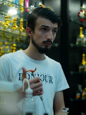 A person is standing indoors in front of shelves filled with colorful, out-of-focus glass containers. They have dark hair and a beard, wearing a white t-shirt with the word 'BONJOUR' visible. They are holding a hookah pipe mouthpiece in one hand.