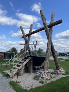 A large wooden structure resembling a swing, with thick logs supporting a suspended metal cauldron over a pile of stones. There are wooden steps leading up to the cauldron. The scene is set outdoors with a grassy area, some trees, and a clear blue sky with scattered clouds.