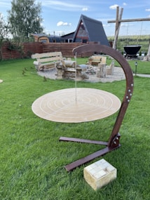 A circular wooden target suspended by a chain from a curved wooden stand is situated on a grassy lawn. In the background, there are log benches, a gravel area with additional wooden seating, and a distinctive triangular-roofed house. The setting suggests an outdoor recreational or relaxation area, with a sky that is partly cloudy.