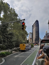 A yellow bus is traveling down a city street lined with trees, with people sitting on an open-top tour bus in the foreground. A city skyline can be seen in the background, including an iconic, tall, triangular building. The traffic light is red, and the street markings indicate a bus lane.