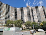 A tall residential building with multiple floors and balconies, set against a clear blue sky. The structure is flanked by lush green trees. At the foreground, a retail space is available for lease, with visible signs advertising it. Several vehicles, including vans and cars, are parked along the street, and a pedestrian walks nearby.