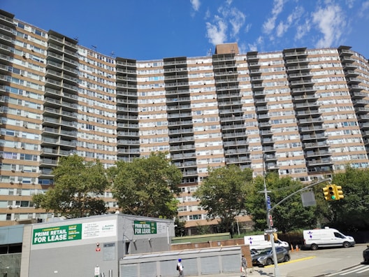 A tall residential building with multiple floors and balconies, set against a clear blue sky. The structure is flanked by lush green trees. At the foreground, a retail space is available for lease, with visible signs advertising it. Several vehicles, including vans and cars, are parked along the street, and a pedestrian walks nearby.