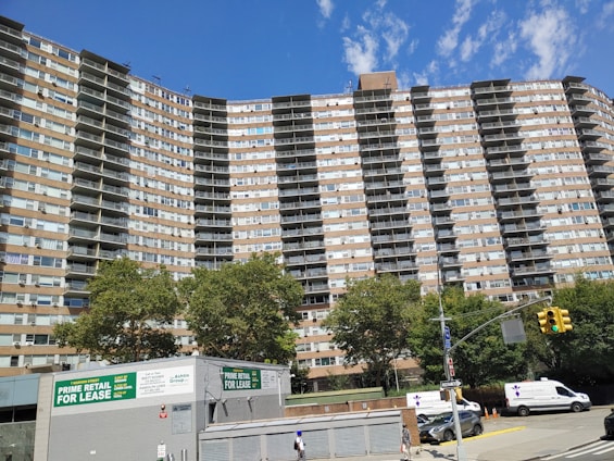A tall residential building with multiple floors and balconies, set against a clear blue sky. The structure is flanked by lush green trees. At the foreground, a retail space is available for lease, with visible signs advertising it. Several vehicles, including vans and cars, are parked along the street, and a pedestrian walks nearby.