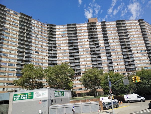 A tall residential building with multiple floors and balconies, set against a clear blue sky. The structure is flanked by lush green trees. At the foreground, a retail space is available for lease, with visible signs advertising it. Several vehicles, including vans and cars, are parked along the street, and a pedestrian walks nearby.