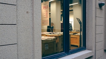 A storefront window displaying a sale sign with prices in a retail store. Inside, wooden tables are arranged with neatly folded clothes, and a person is visible near the back. The store's interior features bright lighting, enhancing the visibility of merchandise indoors.