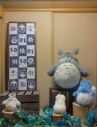 A display of various plush toys resembling characters from a popular animated series is arranged on wooden crates. The large gray plush character sits prominently, surrounded by smaller fluffy toys in white and blue. A framed textile with cartoonish drawings and Asian scripts hangs on the wall to the left.
