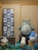 A display of various plush toys resembling characters from a popular animated series is arranged on wooden crates. The large gray plush character sits prominently, surrounded by smaller fluffy toys in white and blue. A framed textile with cartoonish drawings and Asian scripts hangs on the wall to the left.