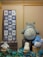 A display of various plush toys resembling characters from a popular animated series is arranged on wooden crates. The large gray plush character sits prominently, surrounded by smaller fluffy toys in white and blue. A framed textile with cartoonish drawings and Asian scripts hangs on the wall to the left.