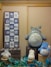 A display of various plush toys resembling characters from a popular animated series is arranged on wooden crates. The large gray plush character sits prominently, surrounded by smaller fluffy toys in white and blue. A framed textile with cartoonish drawings and Asian scripts hangs on the wall to the left.