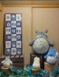 A display of various plush toys resembling characters from a popular animated series is arranged on wooden crates. The large gray plush character sits prominently, surrounded by smaller fluffy toys in white and blue. A framed textile with cartoonish drawings and Asian scripts hangs on the wall to the left.