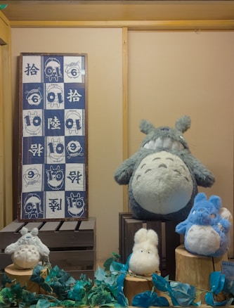 A display of various plush toys resembling characters from a popular animated series is arranged on wooden crates. The large gray plush character sits prominently, surrounded by smaller fluffy toys in white and blue. A framed textile with cartoonish drawings and Asian scripts hangs on the wall to the left.