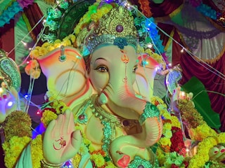 A richly decorated statue of the Hindu deity Ganesha is adorned with elaborate jewelry, a headdress, and surrounded by vibrant garlands of marigold flowers. Colorful lights create a festive atmosphere, illuminating the intricate designs and patterns on the statue. The palm of one hand features an 'Om' symbol.