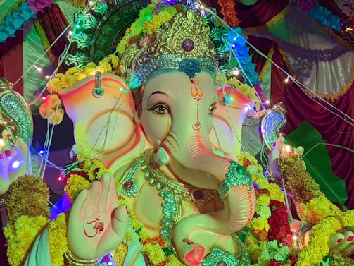 A richly decorated statue of the Hindu deity Ganesha is adorned with elaborate jewelry, a headdress, and surrounded by vibrant garlands of marigold flowers. Colorful lights create a festive atmosphere, illuminating the intricate designs and patterns on the statue. The palm of one hand features an 'Om' symbol.