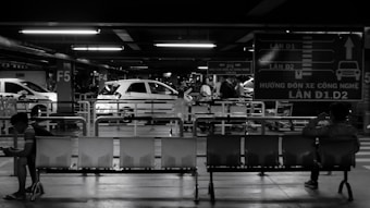 A black and white photograph of a parking garage area is depicted with several people sitting on a row of seats. Cars are parked in the background beneath overhead lighting. Large directional signs are visible, adding to the structured, industrial environment.