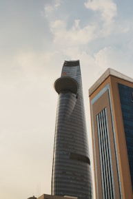 A modern skyscraper with a unique curved design stands prominently against a partly cloudy sky. The building features a circular platform near the top, and its surface is covered in reflective glass. Another high-rise building with blue-tinted windows and brown framing is visible in the foreground.