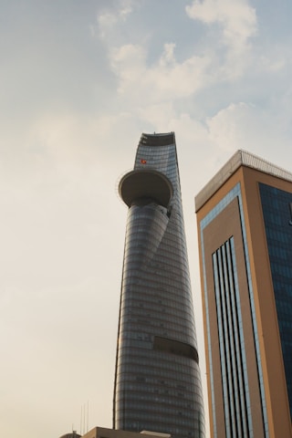 A modern skyscraper with a unique curved design stands prominently against a partly cloudy sky. The building features a circular platform near the top, and its surface is covered in reflective glass. Another high-rise building with blue-tinted windows and brown framing is visible in the foreground.