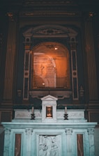An ornate altar is depicted, featuring intricate carvings and a central tabernacle labeled 'MYSTERIUM FIDEI'. The altar is adorned with classical architectural elements, such as columns and carvings of religious figures. A framed artwork or reflective surface hangs above, surrounded by decorative molding.