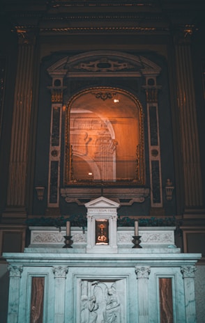 An ornate altar is depicted, featuring intricate carvings and a central tabernacle labeled 'MYSTERIUM FIDEI'. The altar is adorned with classical architectural elements, such as columns and carvings of religious figures. A framed artwork or reflective surface hangs above, surrounded by decorative molding.