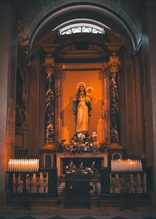 A religious altar featuring a statue of a saint or religious figure holding a child, surrounded by ornate columns and floral decorations. The space is illuminated by numerous lit candles arranged neatly on either side of the altar, creating a warm and solemn atmosphere.