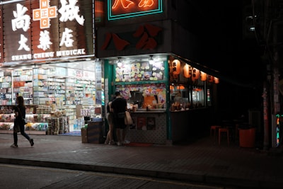 A brightly lit street corner with a medical store on one side and a food stall on the other. The medical store displays a wide range of products visible through large glass windows, with illuminated signage overhead. The food stall is adorned with colorful menus and traditional lanterns, and a few people stand by the counter. Neon signs add vibrant colors to the scene against the nighttime backdrop.