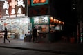 A brightly lit street corner with a medical store on one side and a food stall on the other. The medical store displays a wide range of products visible through large glass windows, with illuminated signage overhead. The food stall is adorned with colorful menus and traditional lanterns, and a few people stand by the counter. Neon signs add vibrant colors to the scene against the nighttime backdrop.