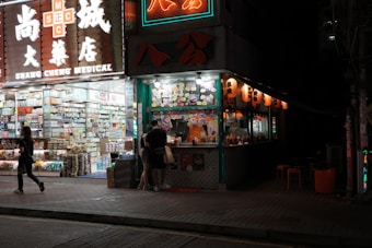 A brightly lit street corner with a medical store on one side and a food stall on the other. The medical store displays a wide range of products visible through large glass windows, with illuminated signage overhead. The food stall is adorned with colorful menus and traditional lanterns, and a few people stand by the counter. Neon signs add vibrant colors to the scene against the nighttime backdrop.