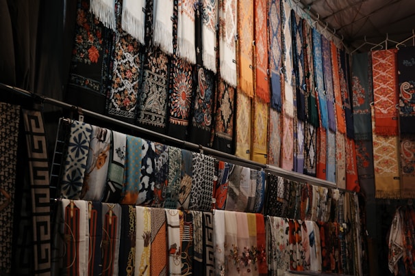 A collection of colorful, intricately patterned fabrics and scarves hangs in rows. The designs include floral patterns, geometric shapes, and other decorative motifs. The fabrics are arranged vertically and appear to be part of a market or shop display.