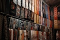 A collection of colorful, intricately patterned fabrics and scarves hangs in rows. The designs include floral patterns, geometric shapes, and other decorative motifs. The fabrics are arranged vertically and appear to be part of a market or shop display.