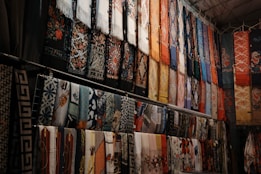 A collection of colorful, intricately patterned fabrics and scarves hangs in rows. The designs include floral patterns, geometric shapes, and other decorative motifs. The fabrics are arranged vertically and appear to be part of a market or shop display.