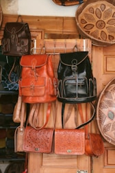 Several handcrafted leather bags and backpacks in various shades of brown and black are hanging on a wooden door. The bags feature intricate patterns and designs. Surrounding the bags are large decorative plates with carved geometric designs.