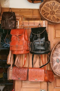 Several handcrafted leather bags and backpacks in various shades of brown and black are hanging on a wooden door. The bags feature intricate patterns and designs. Surrounding the bags are large decorative plates with carved geometric designs.