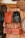 Several handcrafted leather bags and backpacks in various shades of brown and black are hanging on a wooden door. The bags feature intricate patterns and designs. Surrounding the bags are large decorative plates with carved geometric designs.
